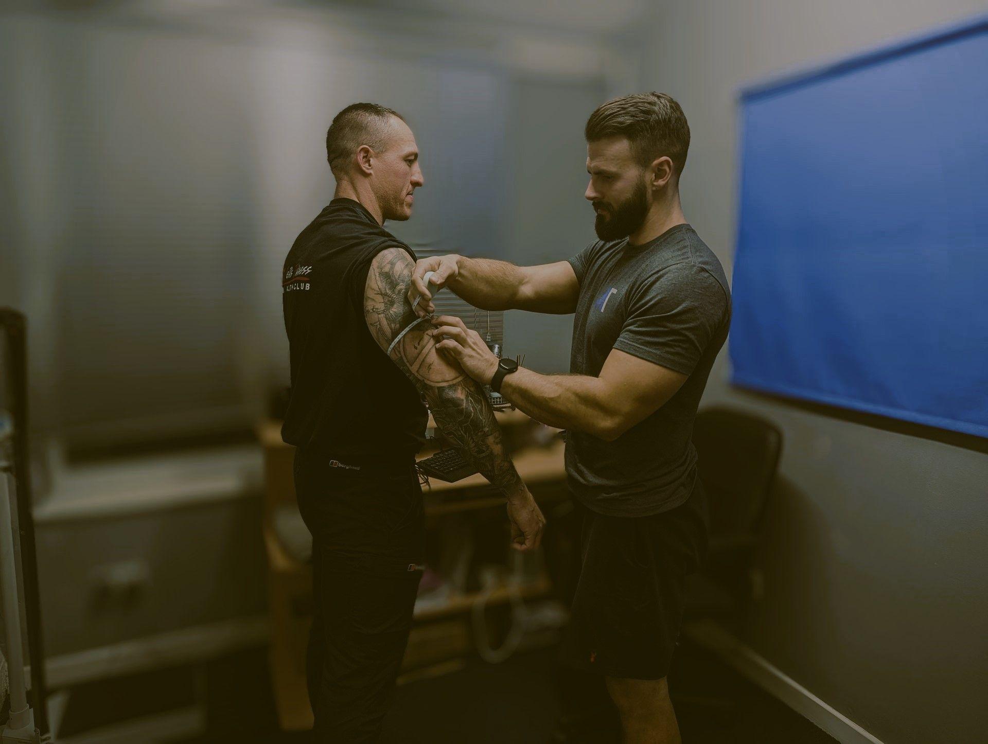 Fitness instructor assisting a client with stretching exercises in a room with gym equipment.