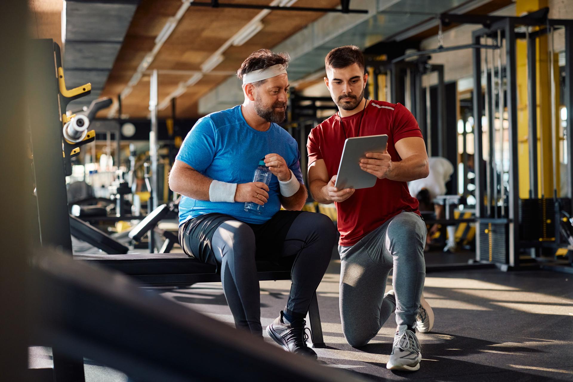 Fitness instructor using touchpad while showing exercise plan to mature athlete in a gym.